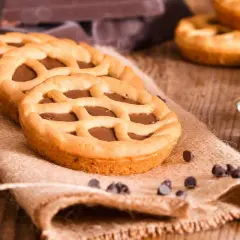 Deliziose crostatine al cioccolato con una griglia di pasta frolla dorata, su un sacco di juta, con gocce di cioccolato sparse.