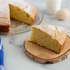 Una fetta di torta 7 vasetti, soffice e cosparsa di zucchero a velo, su un tagliere di legno. Sullo sfondo, la torta intera e un uovo.