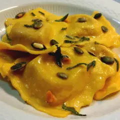 Tortelli di zucca dorati, conditi con erbe aromatiche e semi di zucca tostati, serviti in un piatto bianco.