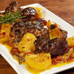 Agnello al forno con patate, succulento e dorato, guarnito con rosmarino fresco su un piatto bianco.