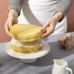 Mani che assemblano un pan di spagna su un'alzata, con ingredienti come uova e burro sul tavolo.