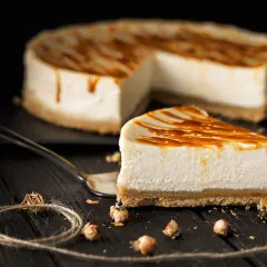 Una fetta di cheesecake senza gelatina, con glassa al caramello, su un piatto scuro. Sullo sfondo, la torta intera.