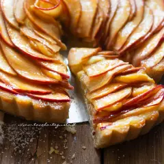 Una deliziosa crostata di mele, con fette di mela lucide disposte a spirale su una base dorata. Una fetta è stata tagliata, rivelando la morbidezza del ripieno.