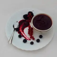 Piatto bianco con salsa ai frutti di bosco, mirtilli, more e ribes. Un cucchiaino d'argento completa la composizione.
