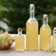 Fiori di sambuco in un cesto e bottiglie di sciroppo fatto in casa. Freschezza estiva da gustare.