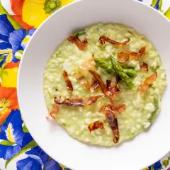 Un cremoso risotto asparagi e speck, guarnito con punte di asparagi freschi e croccanti listarelle di speck, servito su un piatto fiorito.