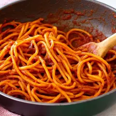 Spaghetti alla sorrentina in una padella, conditi con un ricco sugo rosso e mescolati con un cucchiaio di legno.