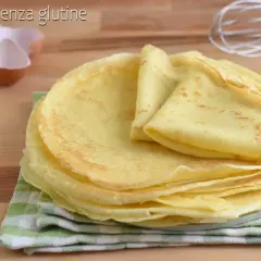 Pila di crepes senza glutine dorate e soffici, pronte per essere farcite. Sullo sfondo, gusci d'uovo e una frusta.