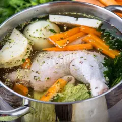 Pentola con brodo di pollo ricetta della nonna: pollo, carote, sedano, cipolla e prezzemolo che sobbollono su un tavolo di legno.