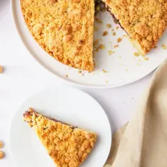 Una fetta di sbriciolata alla Nutella, con la torta intera e ingredienti come nocciole e uova sullo sfondo.