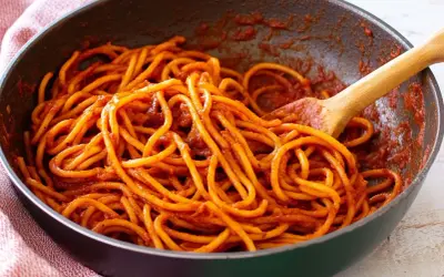 Spaghetti alla sorrentina in una padella, conditi con un ricco sugo rosso e mescolati con un cucchiaio di legno.