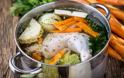 Pentola con brodo di pollo ricetta della nonna: pollo, carote, sedano, cipolla e prezzemolo che sobbollono su un tavolo di legno.