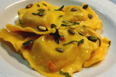 Tortelli di zucca dorati, conditi con erbe aromatiche e semi di zucca tostati, serviti in un piatto bianco.