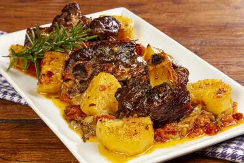 Agnello al forno con patate, succulento e dorato, guarnito con rosmarino fresco su un piatto bianco.