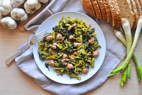 Un piatto di salsiccia e rape saltate, con pane integrale, aglio e cipollotti freschi.