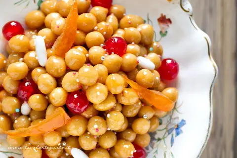 Struffoli ricetta napoletana: palline dorate glassate, ciliegie rosse, canditi e confettini in un piatto decorato.