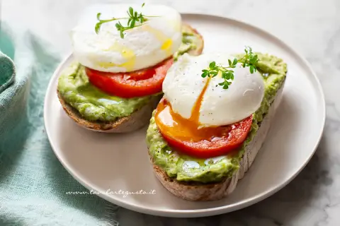Due fette di pane tostato con avocado, pomodoro e un perfetto uovo in camicia, guarnito con germogli freschi.