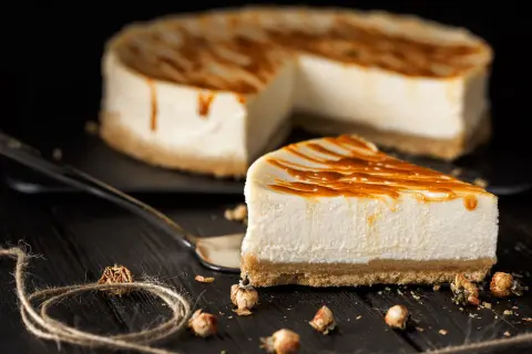 Una fetta di cheesecake senza gelatina, con glassa al caramello, su un piatto scuro. Sullo sfondo, la torta intera.