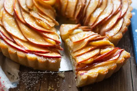Una deliziosa crostata di mele, con fette di mela lucide disposte a spirale su una base dorata. Una fetta è stata tagliata, rivelando la morbidezza del ripieno.
