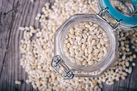 Orzo perlato in un barattolo di vetro, circondato da chicchi sparsi. Attenzione, l'orzo fa ingrassare se consumato in eccesso!