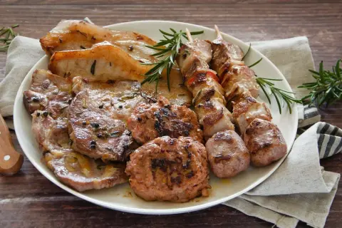 Un piatto ricco di **grigliata di carne**: costine, spiedini, salsicce e hamburger, guarniti con rosmarino fresco.
