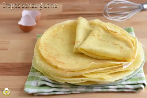 Pila di crepes senza glutine dorate e soffici, pronte per essere farcite. Sullo sfondo, gusci d'uovo e una frusta.