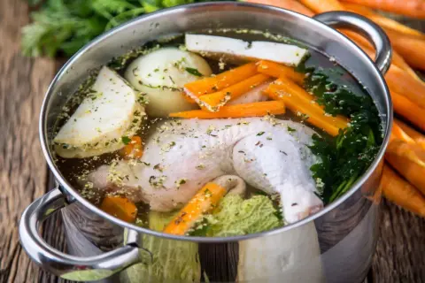 Pentola con brodo di pollo ricetta della nonna: pollo, carote, sedano, cipolla e prezzemolo che sobbollono su un tavolo di legno.