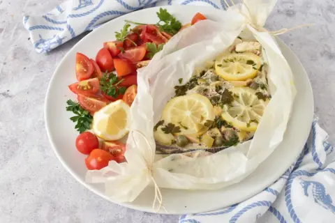 Delizioso pesce spada al forno in cartoccio con limone, capperi e prezzemolo, accompagnato da pomodorini freschi.