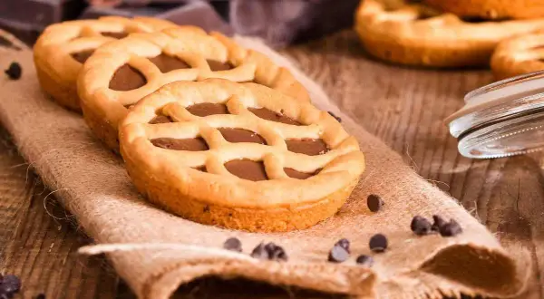 Deliziose crostatine al cioccolato con una griglia di pasta frolla dorata, su un sacco di juta, con gocce di cioccolato sparse.