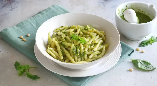 Un piatto invitante di pasta al pesto, condita con basilico fresco e pinoli. Accanto, un mortaio con altro pesto fatto in casa.