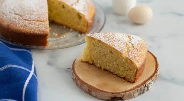Una fetta di torta 7 vasetti, soffice e cosparsa di zucchero a velo, su un tagliere di legno. Sullo sfondo, la torta intera e un uovo.