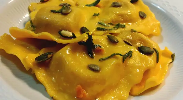 Tortelli di zucca dorati, conditi con erbe aromatiche e semi di zucca tostati, serviti in un piatto bianco.