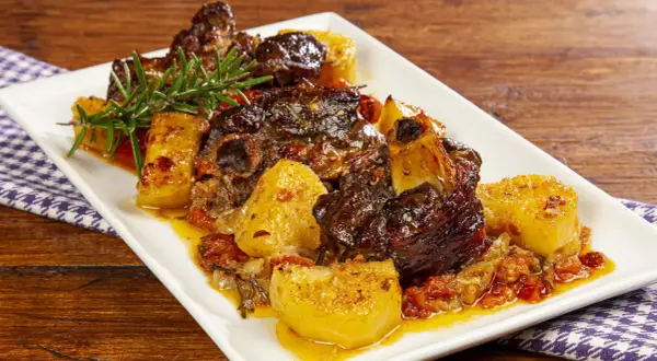 Agnello al forno con patate, succulento e dorato, guarnito con rosmarino fresco su un piatto bianco.