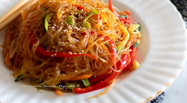 Un piatto invitante di spaghetti di soia con verdure colorate, peperoni rossi, zucchine e carote, guarnito con semi di sesamo.