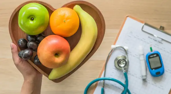Frutta fresca in un vassoio a forma di cuore, accanto a uno stetoscopio e un glucometro, per capire come abbassare la glicemia con una dieta sana.