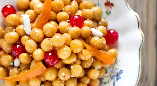 Struffoli ricetta napoletana: palline dorate glassate, ciliegie rosse, canditi e confettini in un piatto decorato.