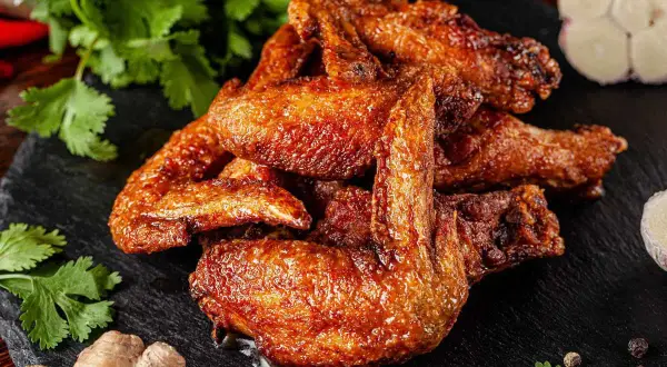Alette di pollo dorate e succulente, servite su una pietra scura con coriandolo fresco, zenzero e aglio.