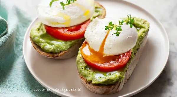 Due fette di pane tostato con avocado, pomodoro e un perfetto uovo in camicia, guarnito con germogli freschi.