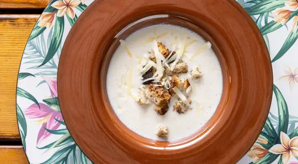 Una cremosa zuppa di cipolle, guarnita con crostini dorati e formaggio grattugiato, servita in una ciotola marrone su un piatto floreale.