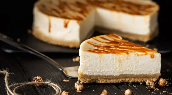 Una fetta di cheesecake senza gelatina, con glassa al caramello, su un piatto scuro. Sullo sfondo, la torta intera.
