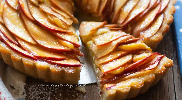 Una deliziosa crostata di mele, con fette di mela lucide disposte a spirale su una base dorata. Una fetta è stata tagliata, rivelando la morbidezza del ripieno.