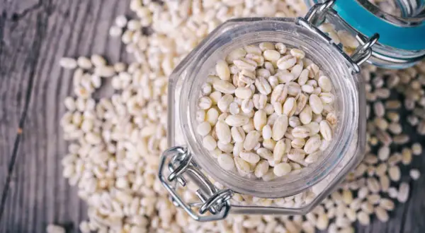 Orzo perlato in un barattolo di vetro, circondato da chicchi sparsi. Attenzione, l'orzo fa ingrassare se consumato in eccesso!