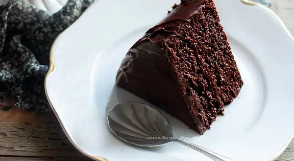 Una fetta di torta al cioccolato, ricca e umida, su un piatto bianco con un cucchiaino.