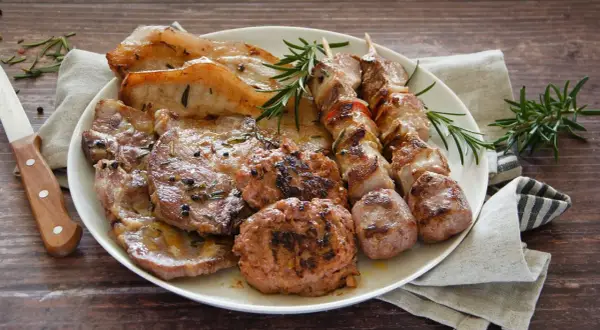 Un piatto ricco di **grigliata di carne**: costine, spiedini, salsicce e hamburger, guarniti con rosmarino fresco.