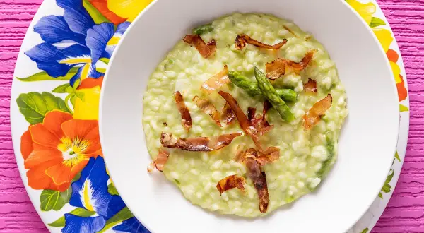 Un cremoso risotto asparagi e speck, guarnito con punte di asparagi freschi e croccanti listarelle di speck, servito su un piatto fiorito.