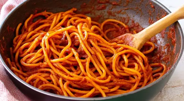 Spaghetti alla sorrentina in una padella, conditi con un ricco sugo rosso e mescolati con un cucchiaio di legno.