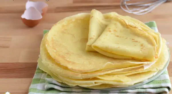 Pila di crepes senza glutine dorate e soffici, pronte per essere farcite. Sullo sfondo, gusci d'uovo e una frusta.