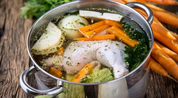 Pentola con brodo di pollo ricetta della nonna: pollo, carote, sedano, cipolla e prezzemolo che sobbollono su un tavolo di legno.