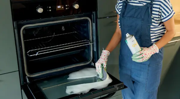 Persona con guanti floreali e salopette in denim pulisce il vetro del forno con una spugna e uno spray.