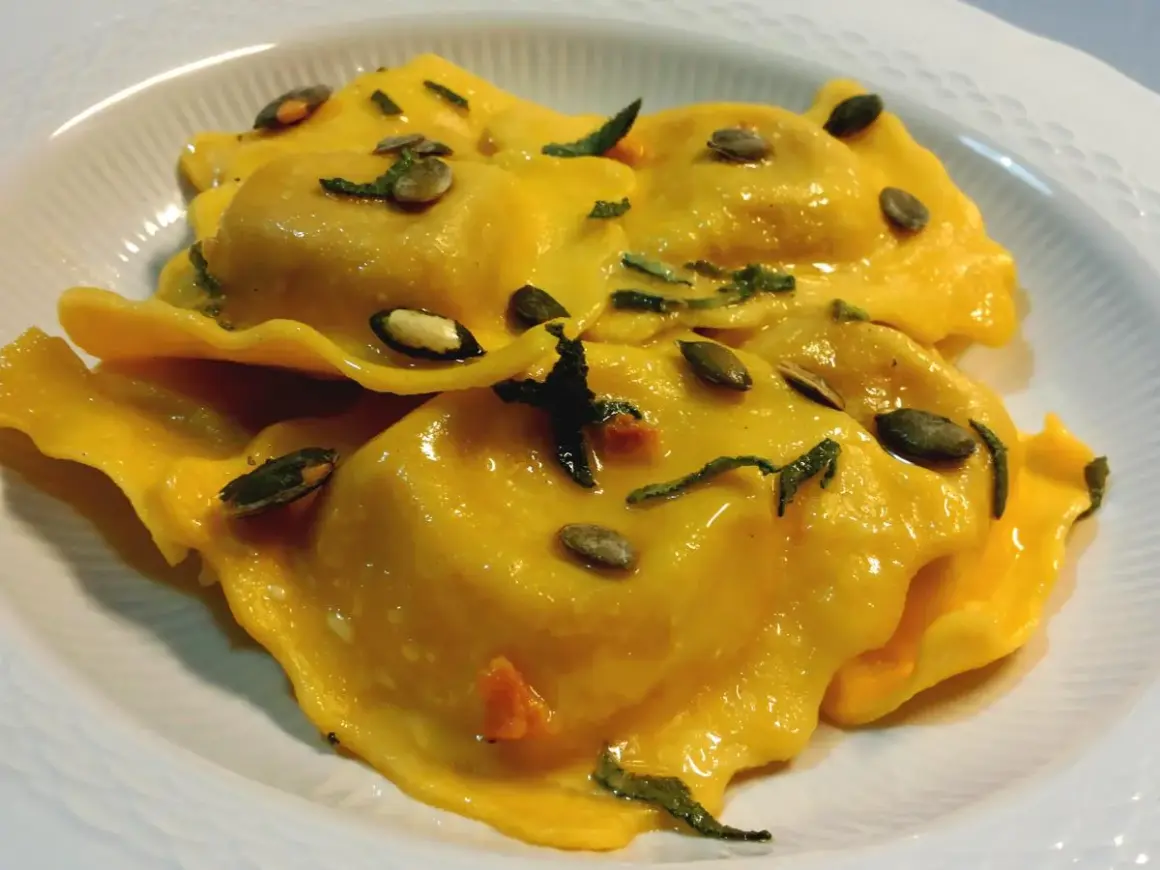 Tortelli di zucca dorati, conditi con erbe aromatiche e semi di zucca tostati, serviti in un piatto bianco.